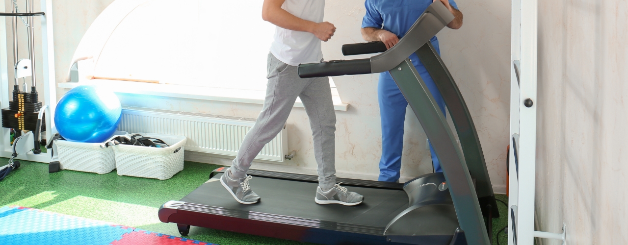 Male physical therapy patient walking on treadmill as physical therapist looks on.